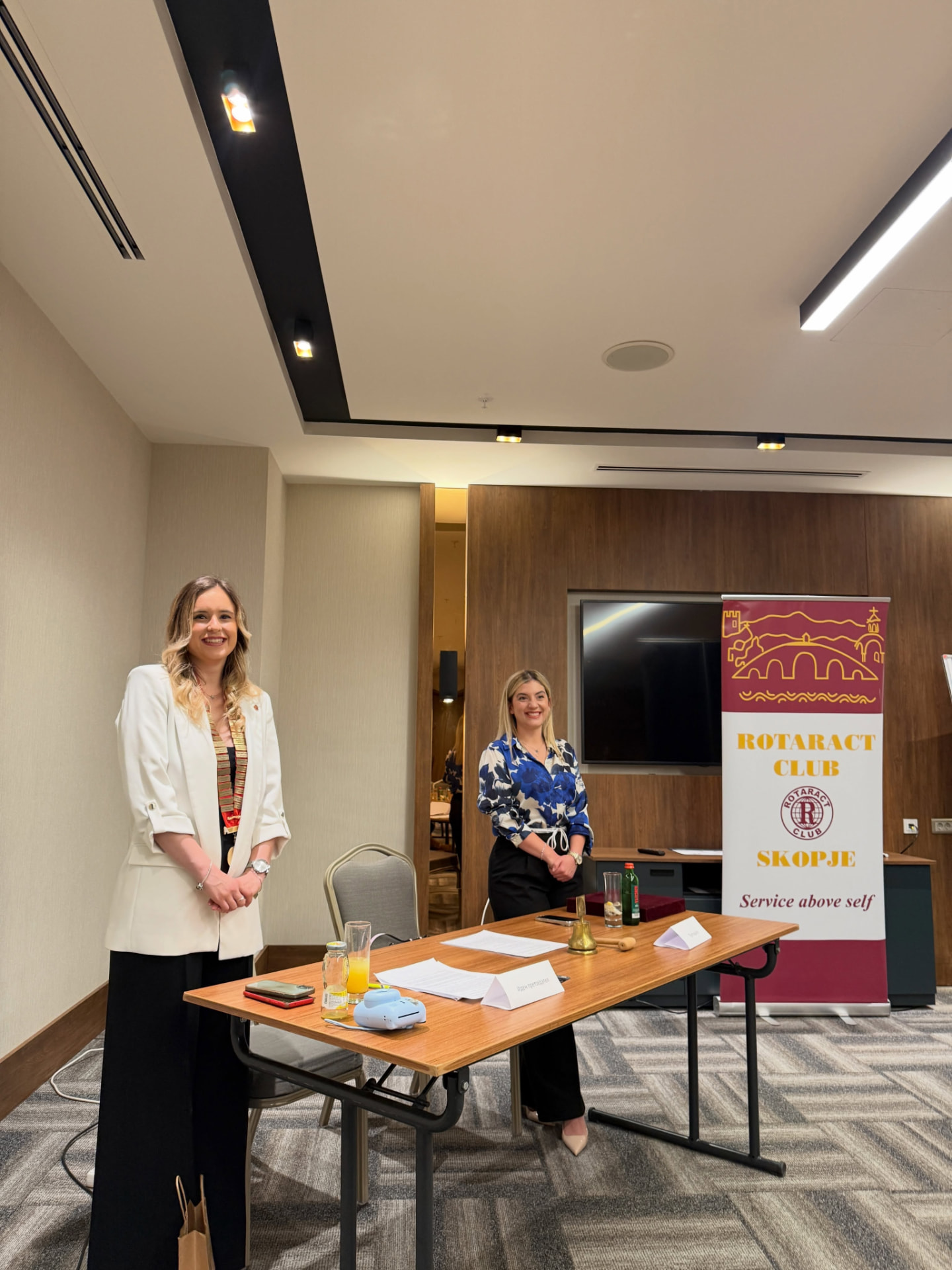 Two members standing at the meeting table with club documents and the bell, next to the Rotaract Club Skopje roll-up banner reading “Service above self”.