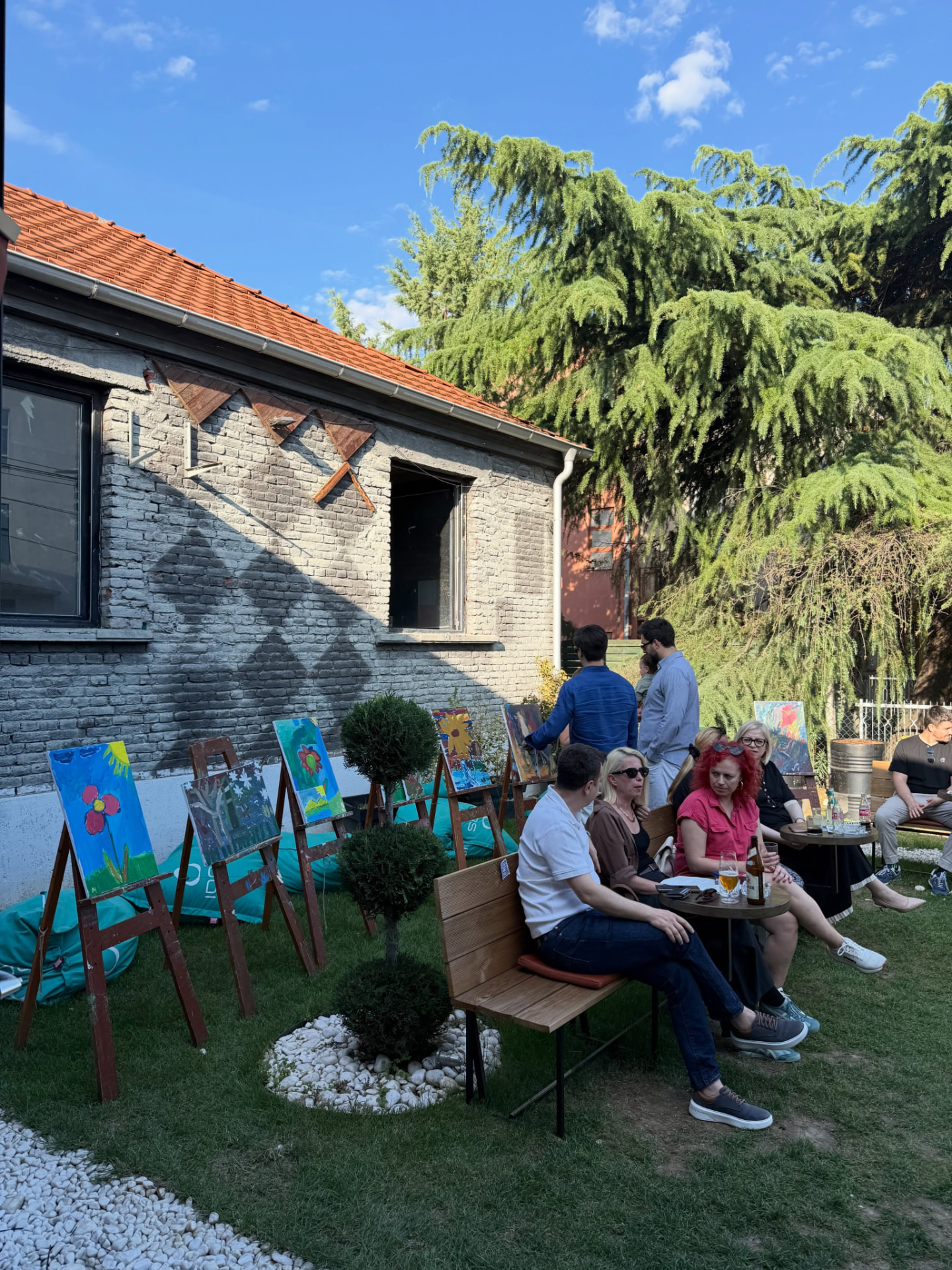 Paintings displayed on wooden easels in a garden while visitors sit at tables nearby on a sunny afternoon.